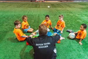 A youth soccer coach in a black jacket that says "Dutch Total Soccer" sitting in a circle with five youth soccer players wearing orange kits